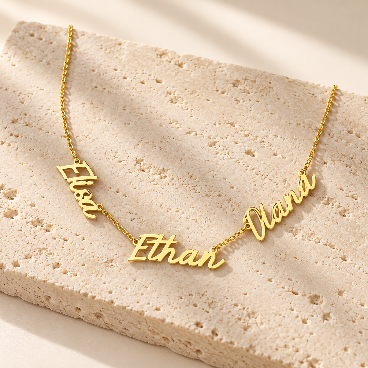Personalized three name necklace in 14K gold vermeil, three script nameplates on a dainty chain, flat lay on travertine stone