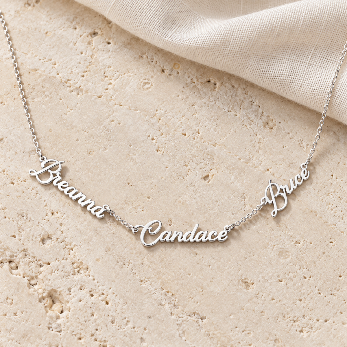 Personalized three name necklace in sterling silver finish, three script names on a cable chain, flat lay on stone